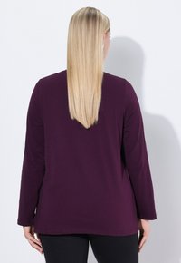 Violetti pitkähihainen paita, jossa on pyöreä pääntie ja löysä istuvuus, valmistettu pehmeästä kankaasta. Helma on hieman pidempi takana.
