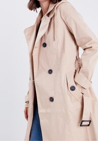 Beige trenchcoat met een dubbelknoopdesign, grote donkere knopen en een verstelbare capuchon. Bevat zijzakken en verstelbare mouwen.