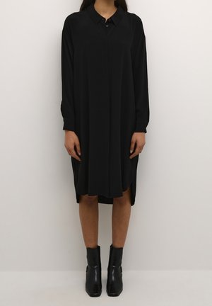 Femme portant une robe midi noire à manches longues avec des boutons et des bottines noires, debout devant un fond clair uni.