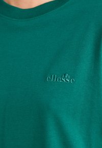 Zelené tričko s okrúhlym výstrihom vyrobené z mäkkej tkaniny. Na ľavej strane má embosované logo v zodpovedajúcej zelenej farbe. Jednoduchý dizajn.