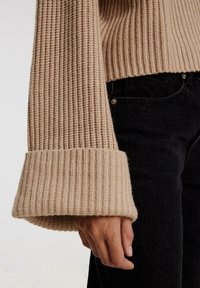 Suéter beige de punto acanalado con puños largos y acampanados. La textura es suave y gruesa, combinado con jeans negros que tienen un detalle de botón plateado.