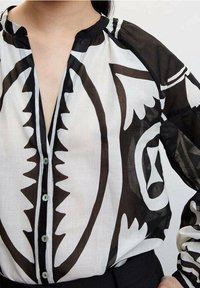 Blusa colletto bianco e nero con audaci motivi astratti, tessuto semitrasparente e chiusura a bottoni sul davanti. Maniche lunghe con accenti a contrasto.