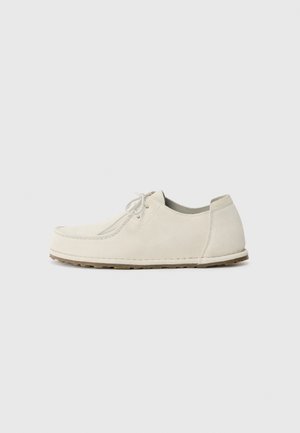 Chaussure en daim beige clair au design arrondi, avec un simple système de laçage et une semelle en caoutchouc texturée.