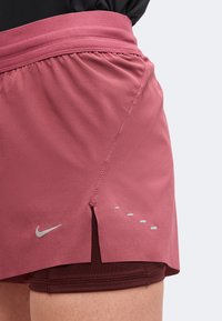 Άτομο που φοράει ροζ σορτς Nike για τρέξιμο με αντανακλαστικές λεπτομέρειες και εσωτερικό μπορντό στρώμα, φαίνεται από τη μέση μέχρι το πάνω μέρος του μηρού.