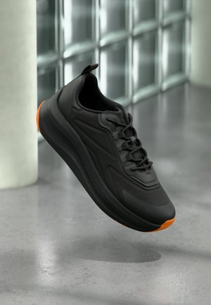 Schwarzer Sportschuh mit orangefarbenen Akzenten, der über einem polierten grauen Boden in einem modernen Innenraum mit Glasbausteinen schwebt.