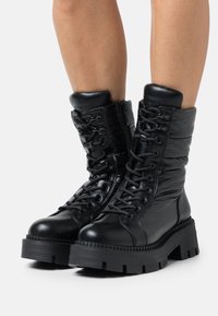 Botas negras hasta el tobillo con una combinación de cuero y material acolchado. Presenta una suela gruesa, cordones y un cierre de cremallera en el lateral.