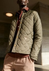 Vadderad olivgrön jacka med mässingsknappar, med en bröstficka. Bärs över en vinröd skjorta, kombinerad med ribbade rosa byxor.