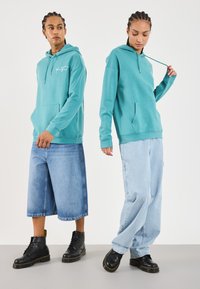 Sudadera con capucha de color turquesa y bolsillo frontal, combinada con vaqueros de pierna ancha de color azul claro. Ambos llevan botas de cordones negras con acentos amarillos.