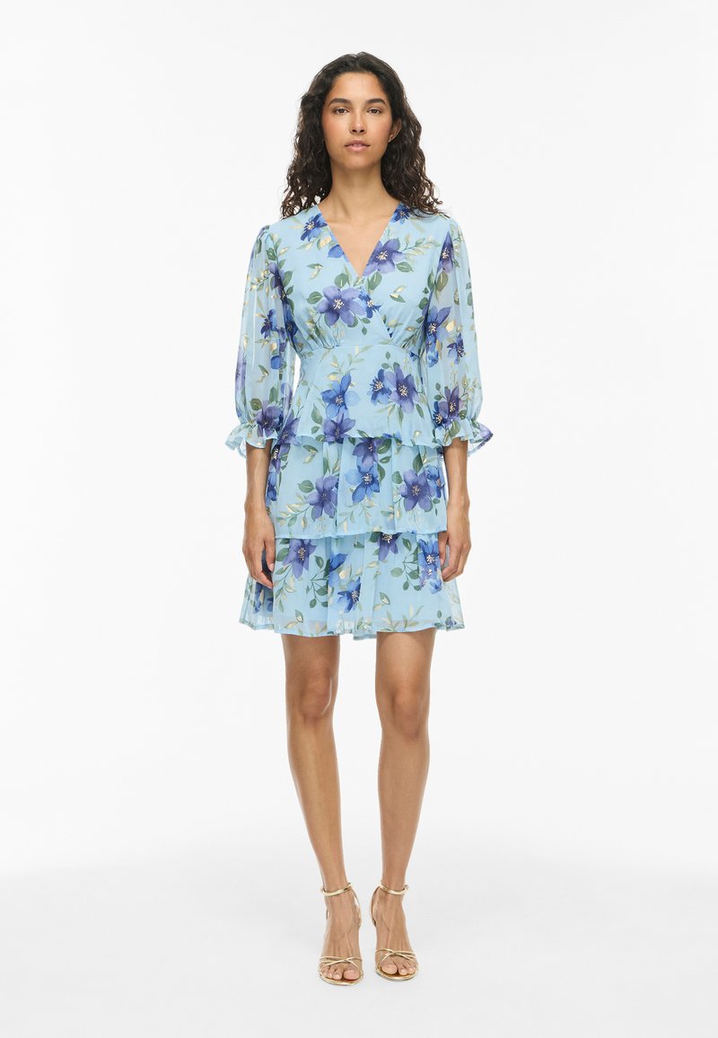 Femme portant une robe portefeuille fleurie bleu clair avec manches trois-quarts et jupe à volants, debout et faisant face à l'avant sur fond blanc.