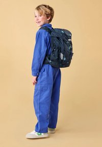 Zaino in tessuto scuro con motivo stellato, spallacci imbottiti e tasche laterali in rete. Il modello indossa una salopette blu e scarpe bianche con dettagli verdi.