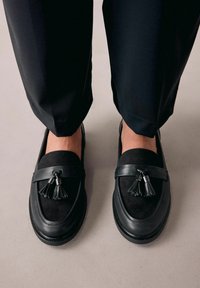 Sorte loafers i en kombination af glat læder og blødt ruskind, med dekorative kwast og en tekstureret gummisål.