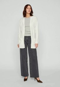 Cardigã longo branco com frente aberta, textura canelada e dois bolsos frontais, combinado com uma blusa cinza e jeans escuros de perna larga. Sapatos de salto castanhos.