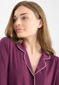 Top de pijama con botón automático y escote en V profundo en un rico tejido morado, que presenta un ribete blanco contrastante en el cuello y los puños. Textura suave.