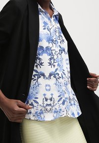 Camicia a fiori bianca con motivi blu. Presenta maniche corte e colletto. Indossata sotto un blazer nero e abbinata a una gonna leggera verde chiaro con una texture.