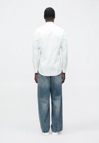 Fehér, hosszú ujjú, egyenes szabású ing, laza, világoskék farmernadrággal párosítva. A ruházaton egyszerű gombos részletek találhatók.