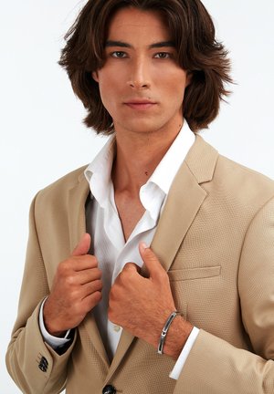 Beige kavaj över vit skjorta, modell som håller i kragen, bär en silverarmband, slätt hår, neutral bakgrund.