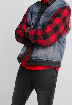 Person iført en rød og sort ternet flannel-skjorte under en blå denimjakke med sort kant, armene krydsede, og mørke jeans.