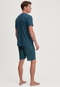 T-shirt en coton bleu-vert associé à un short foncé à motifs avec des formes géométriques blanches. Le modèle se tient pieds nus, vu de dos.