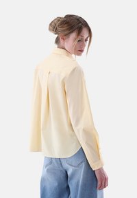 Light yellow long-sleeve shirt with a classic collar, back pleat, and buttoned cuffs, paired with loose blue denim trousers.