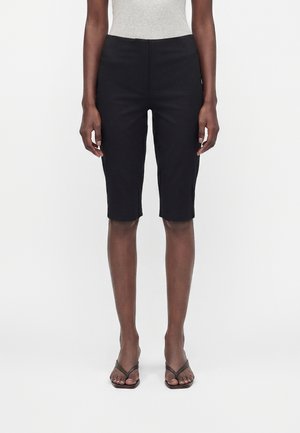 Personne portant un short noir jusqu'aux genoux, un débardeur gris et des sandales plates marron, debout devant un fond blanc.