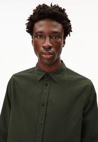 Young man with short curly hair wearing a dark green button-up shirt, facing forward with a neutral expression on a white background.