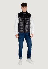Chaleco de plumas negro con textura brillante, cierre de cremallera y cuello alto sobre una camiseta blanca de manga larga. Combinado con jeans azules y zapatillas blancas.