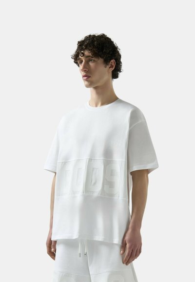 Giovane uomo con capelli ricci che indossa una maglietta bianca larga e pantaloncini bianchi abbinati, su uno sfondo bianco uniforme.