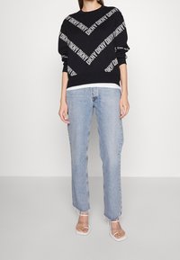Schwarzer Pullover mit weißem "DKNY"-Logo, Rundhalsausschnitt, lange Ärmel, kombiniert mit hellblauen, geraden Jeans und durchsichtigen Abendsandalen.