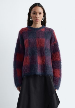 Femme portant un pull à carreaux flou rouge et bleu, une jupe asymétrique noire, des chaussures noires, et portant un sac à bandoulière noir texturé.