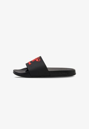 Sandalia slide negra con una correa ancha que presenta un logotipo de abeja estilizado en rojo y el nombre de la marca, suela plana, vista lateral.
