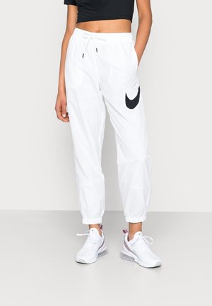 Person trägt weiße Nike-Sweathosen mit schwarzem Logo, weiße Sneakers und ein schwarzes kurzes Top, steht vor einem schlichten Hintergrund.