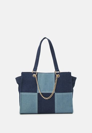 Tote-taske med patchwork af mørk og lyseblå denimpaneler, dobbelte stofhåndtag og en guldkæde-detalje på en hvid baggrund.