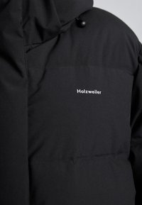 Fekete puffernadrág kapucnival, sima szövet textúrával és egy kis fehér "Holzweiler" logóval a mellkason.