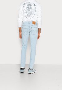 Jasnoniebieskie jeansy o dopasowanym kroju z skórzaną metką na pasku. Połączone z białą bluzką z długim rękawem z graficznym wzorem.