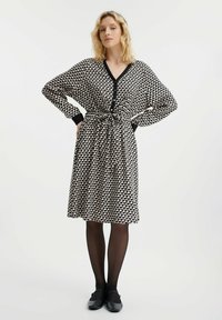 Czarna sukienka w kremowy wzór z dekoltem w kształcie litery V, długimi rękawami i zapięciem na guziki, z przewiązanym w pasie paskiem i luźnym krojem, zestawiona z ciemnymi rajstopami.