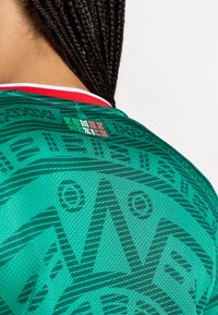 Camisa de desporto verde com mangas curtas, apresentando um padrão texturizado e uma gola vermelha. Um emblema está posicionado nas costas.
