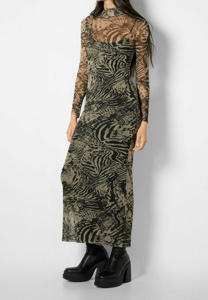 Mujer con vestido largo, transparente y de cuello alto con patrón abstracto en negro y verde oliva y botines negros con plataforma y tacón.