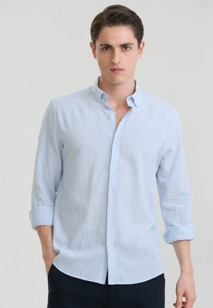 Junger Mann mit kurzen braunen Haaren, der ein hellblaues, vertikal gestreiftes Langarmhemd mit hochgekrempelten Ärmeln und dunkle Hosen trägt.