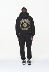 Černá mikina s kapucí se velkým žlutým logem "PEGADOR" a textem "BUILT TO LAST" na zadní straně, doplněná o odpovídající černé teplákové kalhoty.