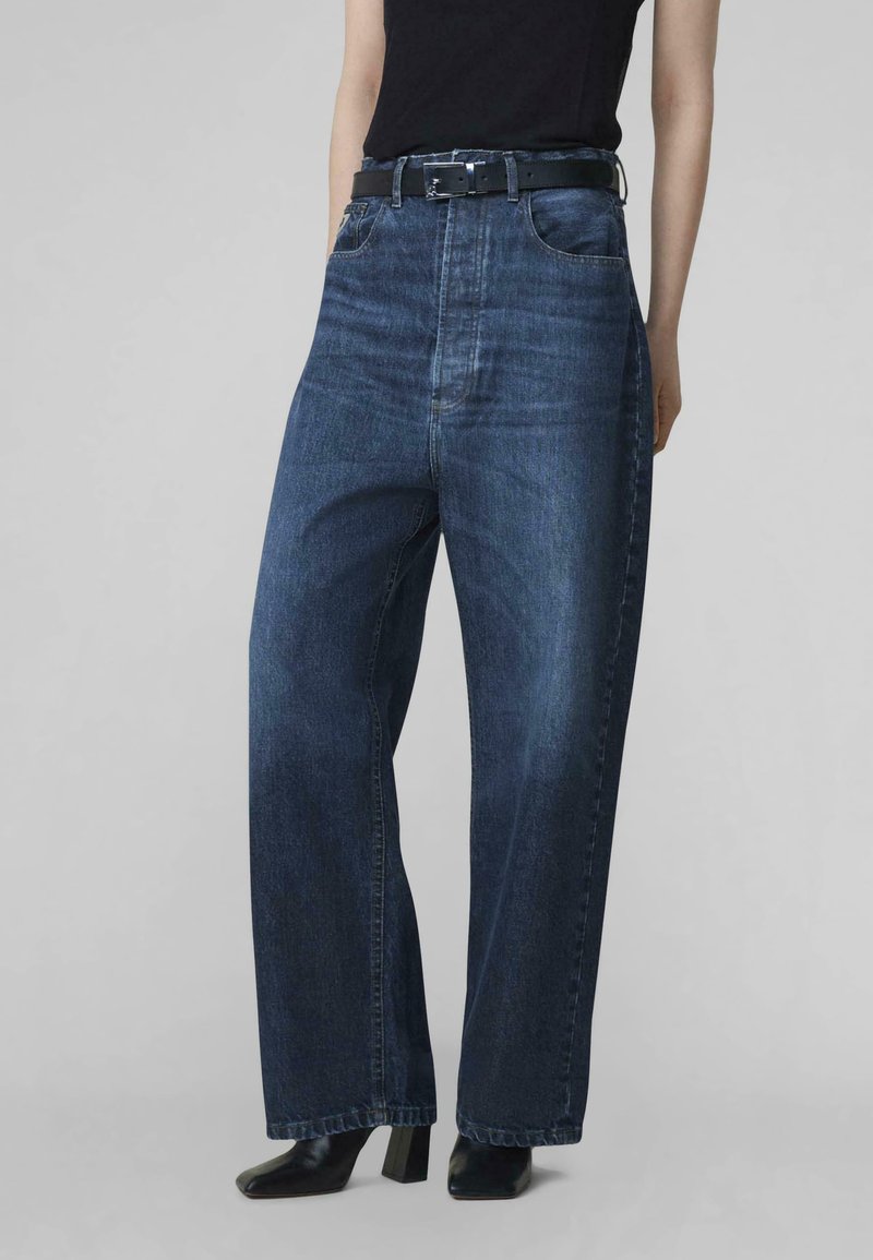 Jeans de mezclilla oscura de pierna ancha con cintura alta, trabillas para cinturón, bolsillos frontales y un sutil desgaste, combinados con un cinturón negro y botas de tacón.