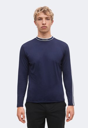 Ung mand med kort krøllet blondt hår iført en navyblå langærmet skjorte med hvide striber og et Calvin Klein-logo på kraven.