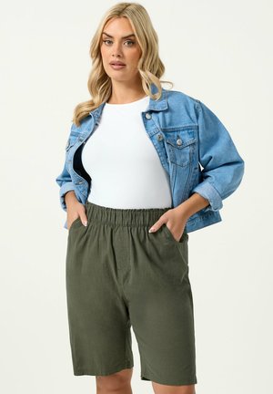 Femme blonde portant une veste en jean bleue sur un haut blanc et un short taille haute vert olive, les mains dans les poches, debout devant un fond blanc.