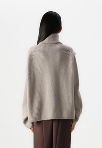 Un pull surdimensionné à côtes de couleur gris clair avec un col montant, présentant des épaules tombantes et une texture confortable. Assorti à un pantalon marron.