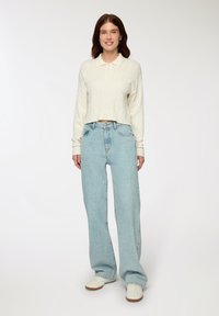 Donna in piedi che indossa un maglione a polo in crema con lavorazione a trecce, jeans a gamba larga blu chiaro e sneakers bianche su uno sfondo neutro.