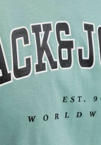 Lettres majuscules noires en gras "JACK&J" avec un texte plus petit "EST. 9" et "WORLDW" sur un fond de tissu vert clair.