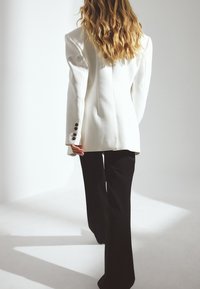 Bela strukturirana blejzer s podloženimi rameni, v kombinaciji s črnimi širokimi hlačami. Vsebuje črne gumbe na rokavih. Teksturni material.