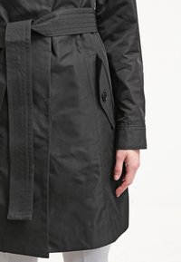Manteau noir ceinturé avec poche à rabat boutonnée et manches longues porté par une personne sur un fond uni.