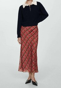 Pull zippé bleu marine avec col crème, associé à une jupe à carreaux rouges avec des lignes roses et marron, porté avec des ballerines noires pointues.