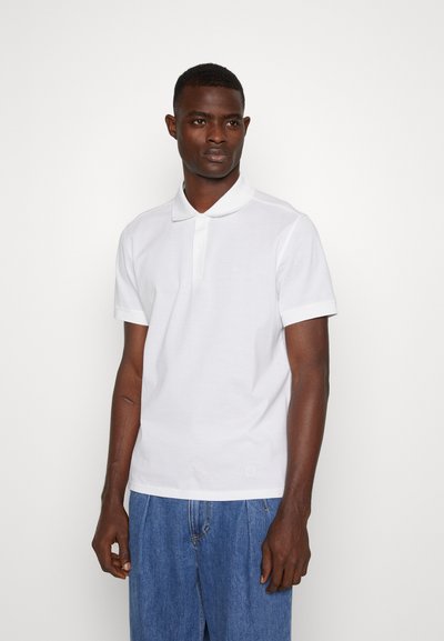 Camisa polo blanca de manga corta, con cuello clásico y una sutil textura en la tela. Combinada con pantalones cortos de mezclilla azul claro.