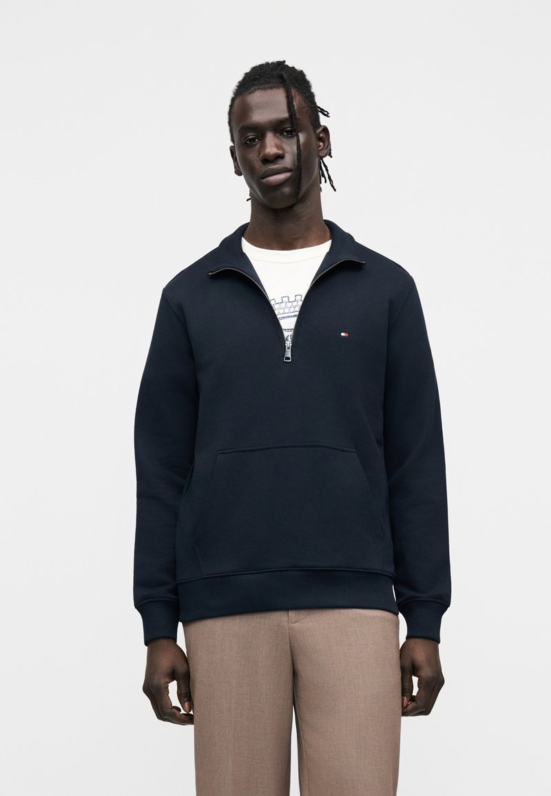 Μπλε ναυτικό φούτερ με φερμουάρ, ψηλό γιακά, τσέπη καγκουρό και μικρό λογότυπο στο στήθος. Το ύφασμα έχει λεία, απαλή υφή.